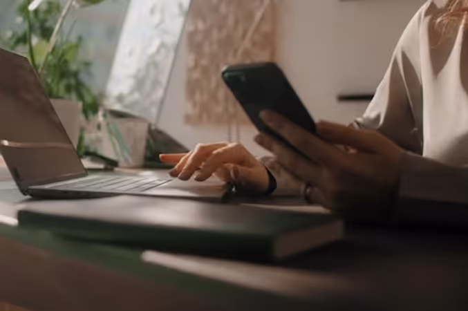 Person using a smartphone with one hand while typing on a laptop keyboard with the other on a desk.