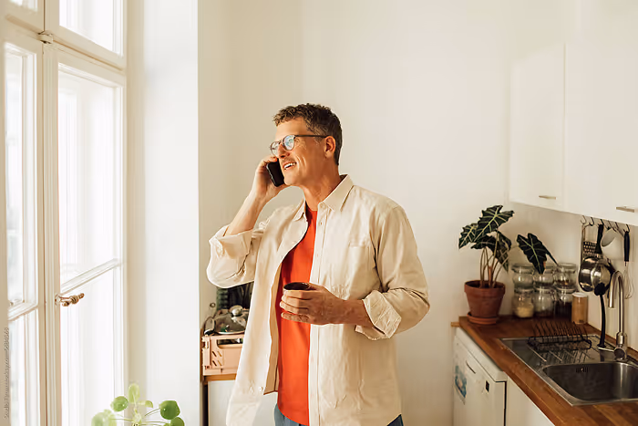 Man with glasses standing in a kitchen, smiling and talking on a phone while holding a coffee cup.