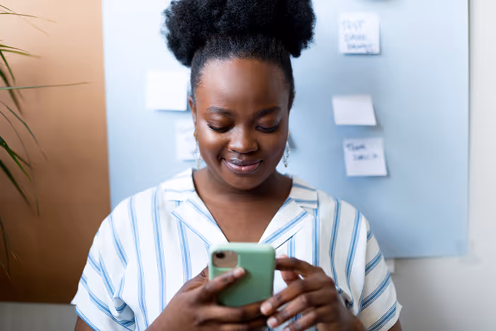 Smiling woman looking at her smartphone, standing indoors with sticky notes on a board behind her.