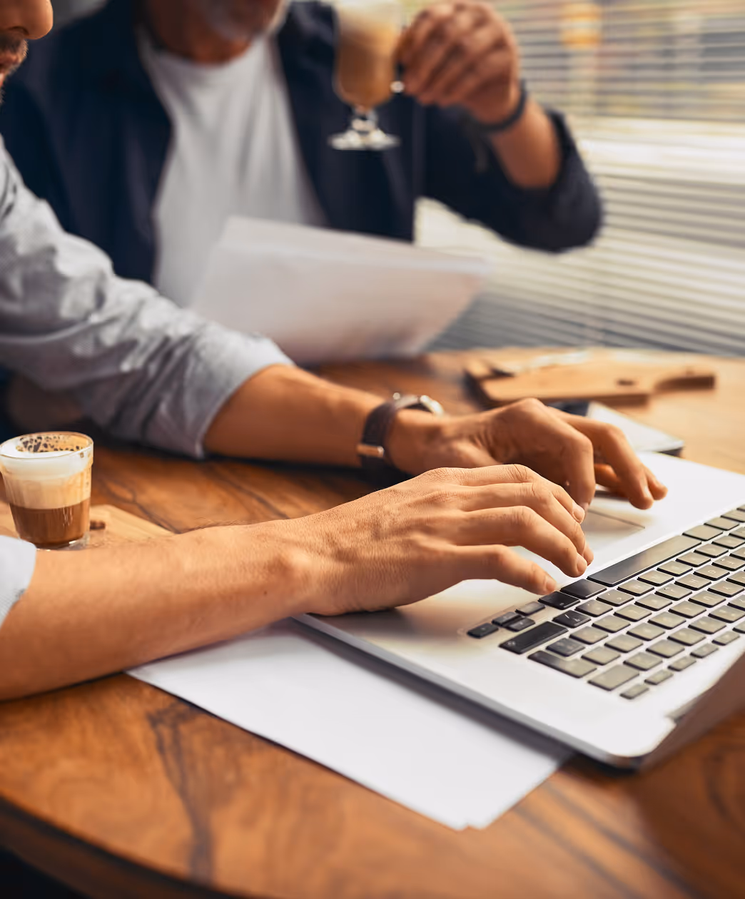 Close-up of two men at a wooden table working on a laptop with coffee and documents nearby.