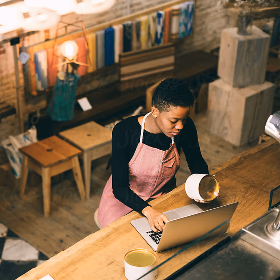 Barista in a pink apron working on a laptop behind a wooden counter in a coffee shop.