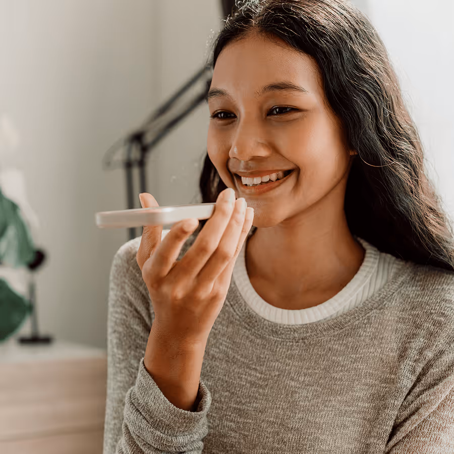 Smiling woman holding a smartphone close to her mouth, speaking into it indoors.