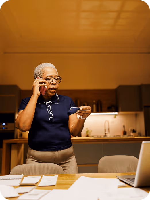 Elderly woman in glasses talking on the phone while holding a credit card in a kitchen.