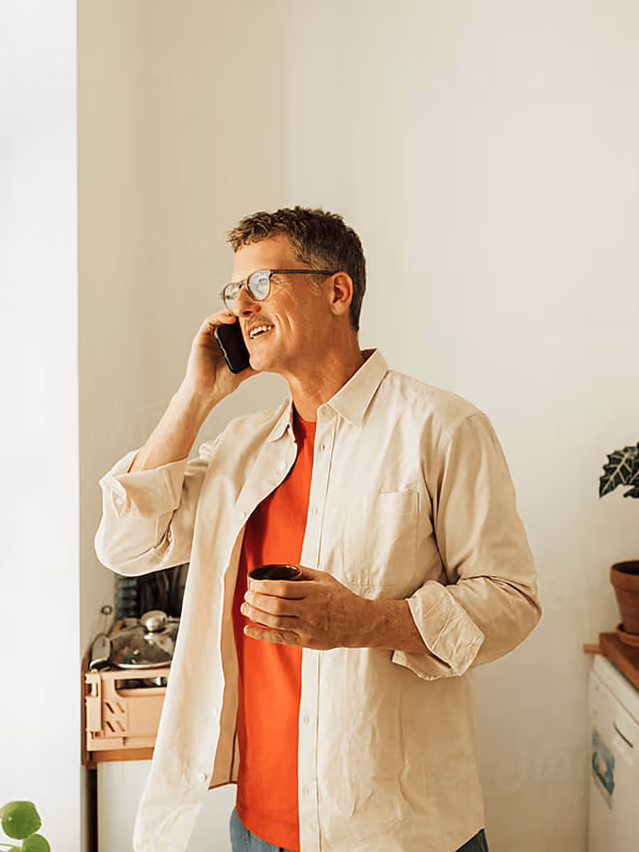 Middle-aged man wearing glasses and a cream shirt holding a coffee cup while talking on a smartphone indoors.