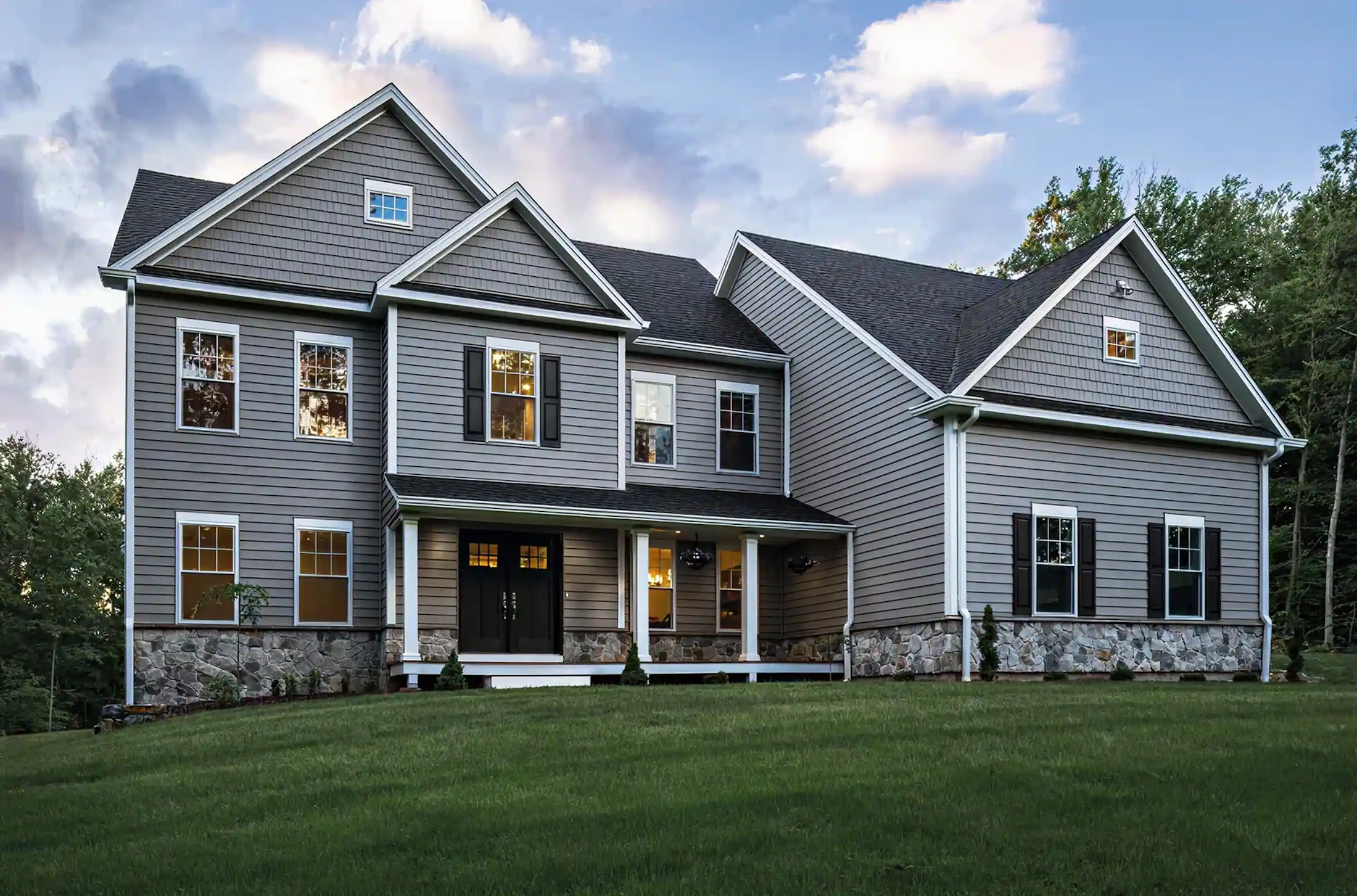 Two-story gray house with stone foundation, black shutters, white-trimmed windows, and a covered porch on a green lawn.