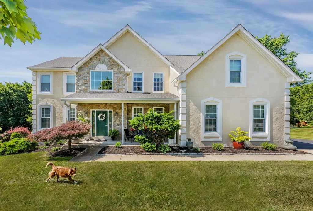 Two-story beige house with a green front door, stone accents, landscaped yard, and a brown dog running on the lawn.