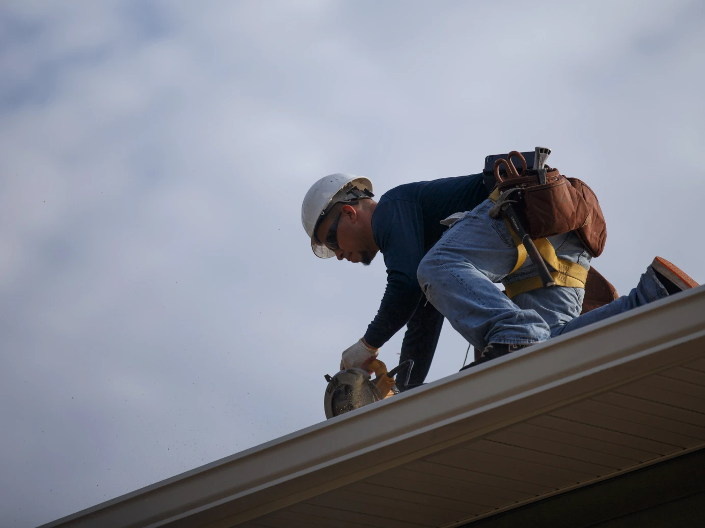 Construction worker wearing a white hard hat and safety harness using a circular saw on a roof under a cloudy sky.