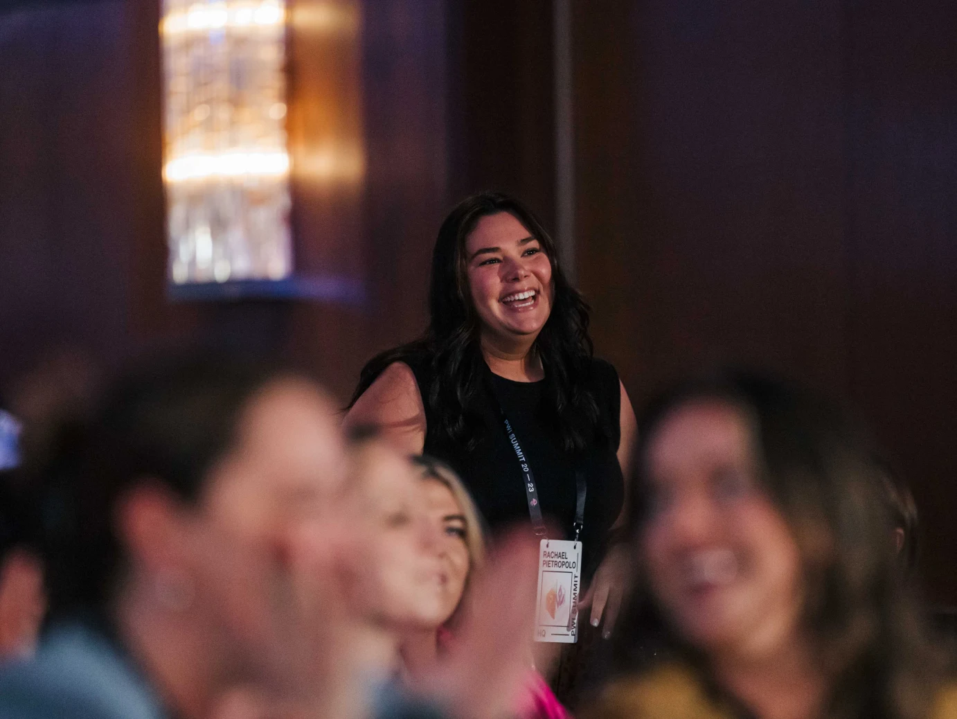Smiling woman with dark hair wearing a black sleeveless top and event badge in a dimly lit room.
