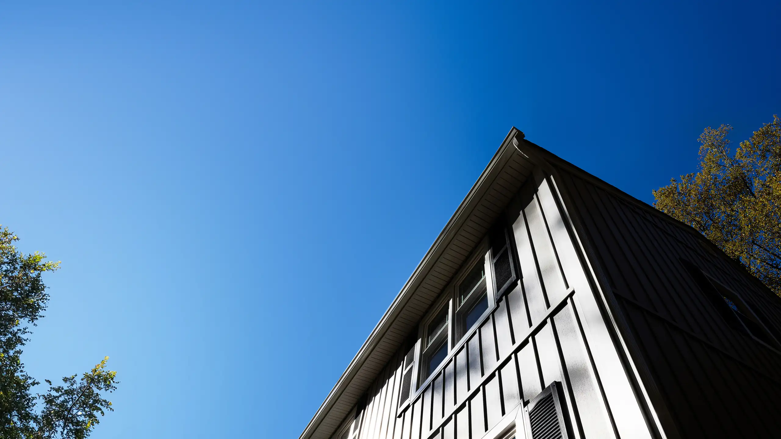 Upward view of a white pitched-roof house with large windows and siding against a clear blue sky with trees on the sides.