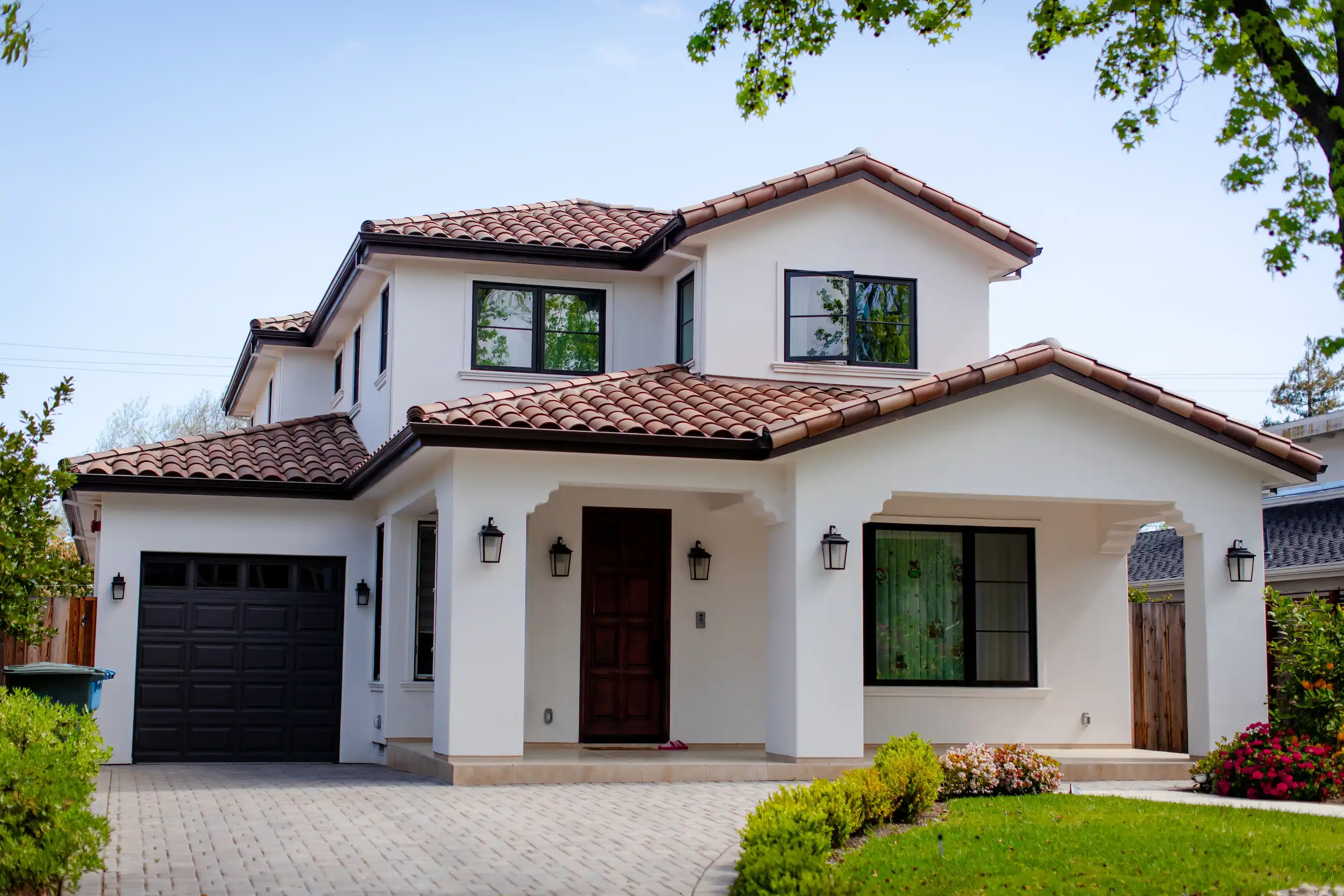 Two-story white house with a red-tiled roof, dark garage door, wooden front door, and landscaped front yard.