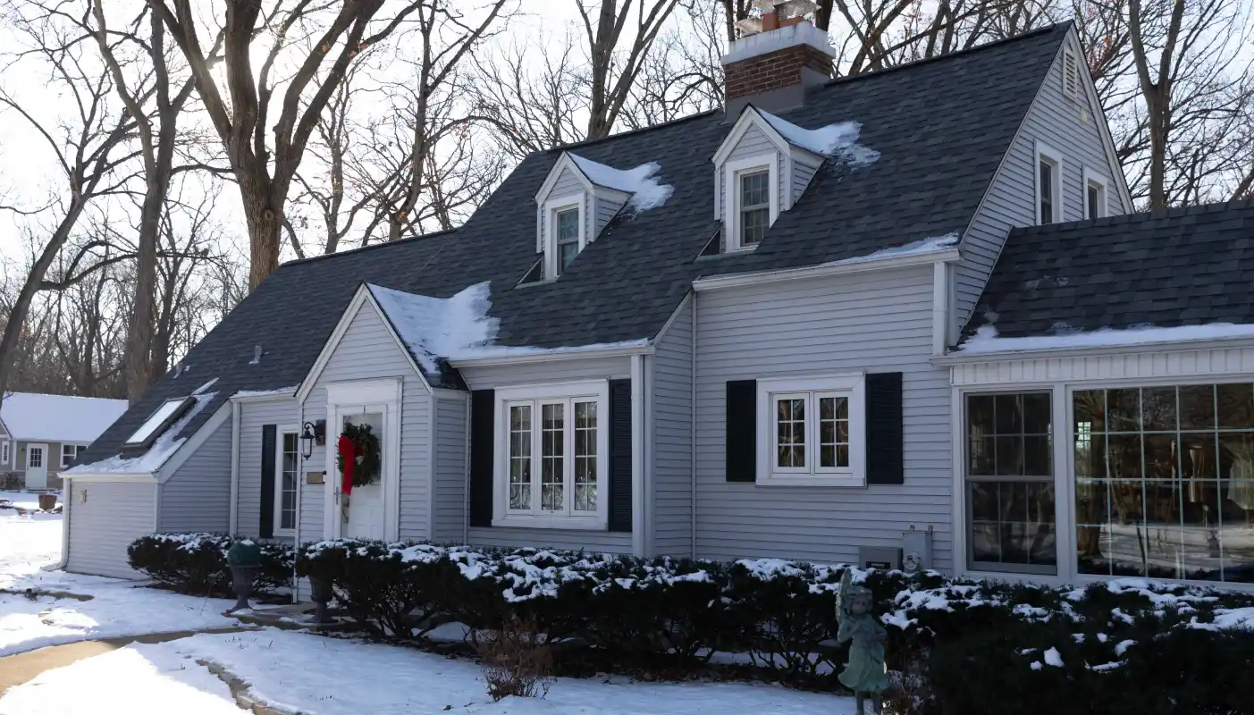 Gray house with steep roof and partial snow cover on roof and ground, decorated with a Christmas wreath on the front door, surrounded by leafless trees.