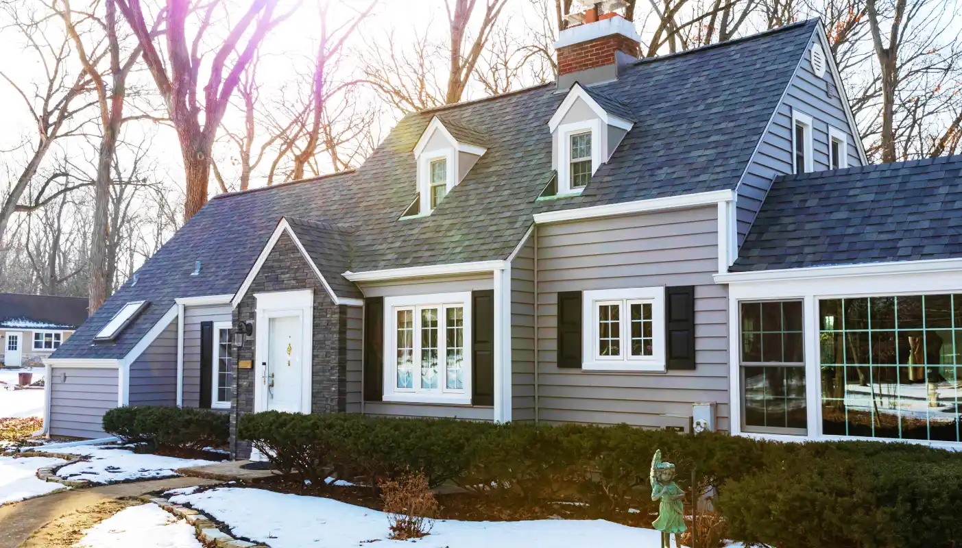 Gray house with a white door, dormer windows, and surrounding snow-covered yard with bushes and a small statue.