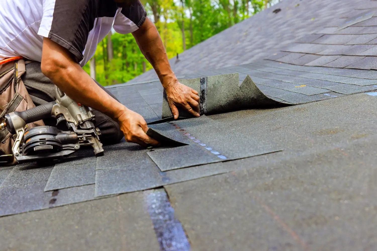 A worker installing gray asphalt shingles on a roof with a nail gun nearby and green trees in the background.