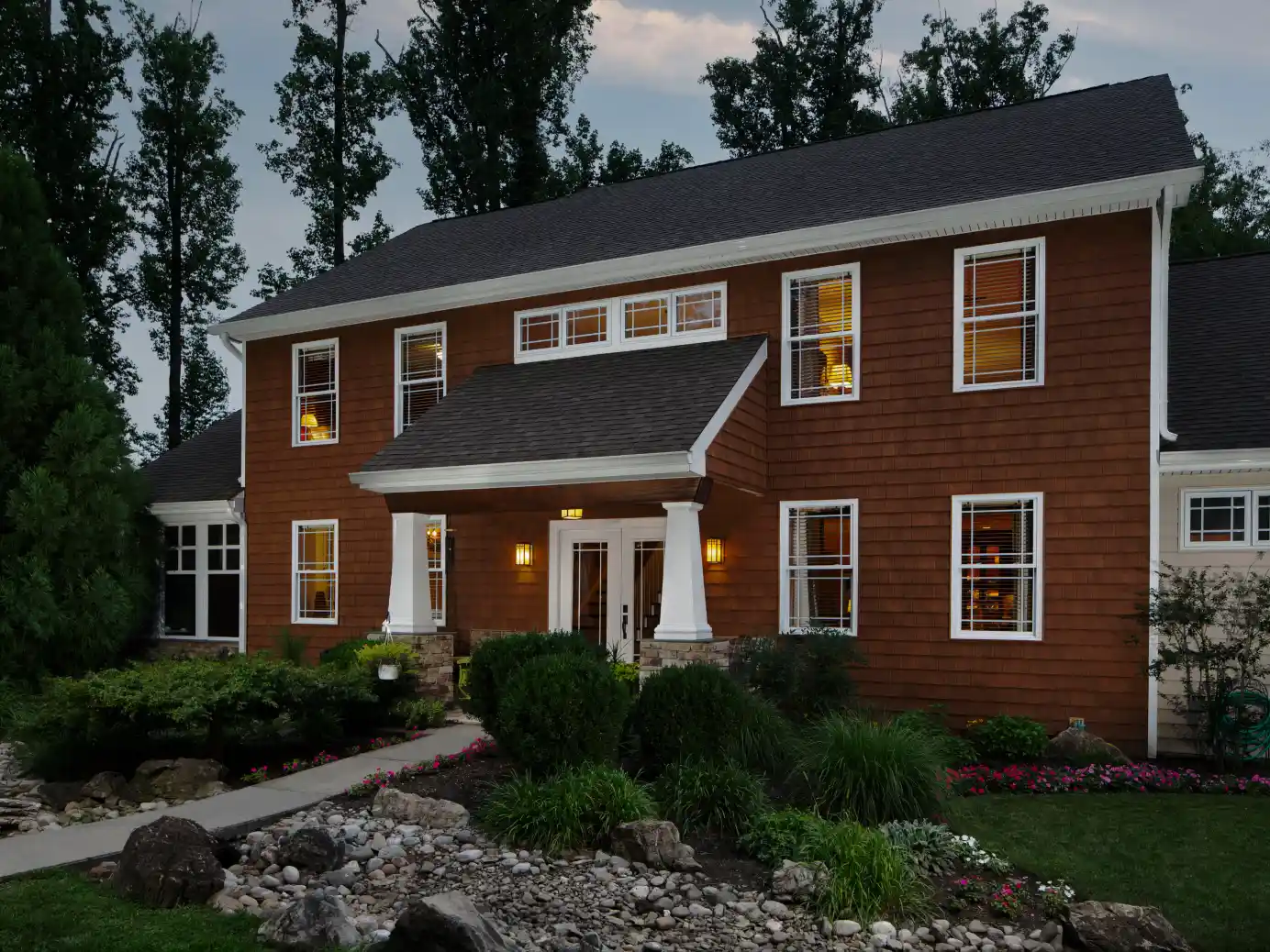 Two-story house with brown siding, white trim, front porch with pillars, lit windows, and landscaped garden with rocks and bushes at dusk.