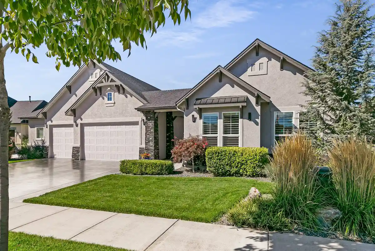 Modern single-story house with three-car garage, manicured lawn, shrubs, and trees under a blue sky.
