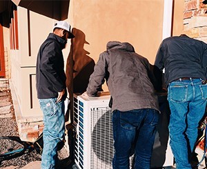 Three men inspecting or repairing an outdoor air conditioning unit next to a building wall.
