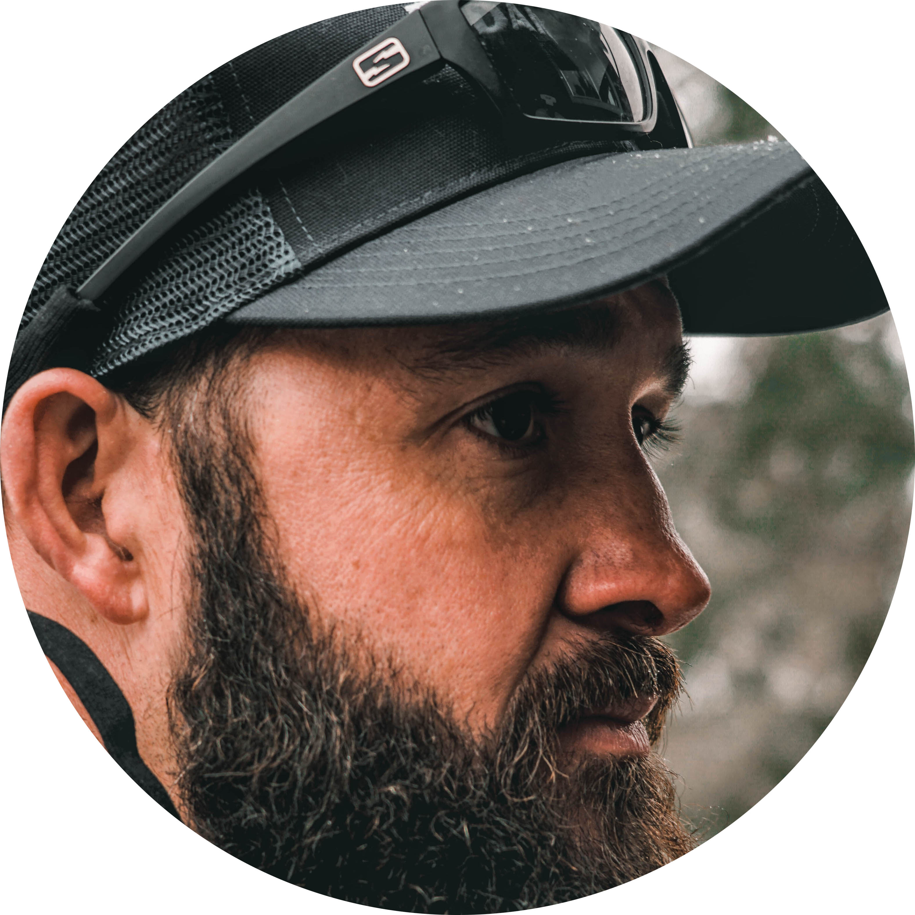 Side profile of a bearded man wearing a black mesh cap and sunglasses on the cap.