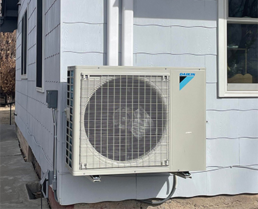 Daikin brand air conditioning unit mounted on the exterior wall of a light blue house under a window.