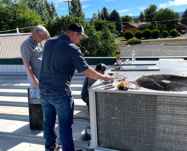 Two men working on rooftop HVAC unit with tools on the unit's surface under a sunny sky.