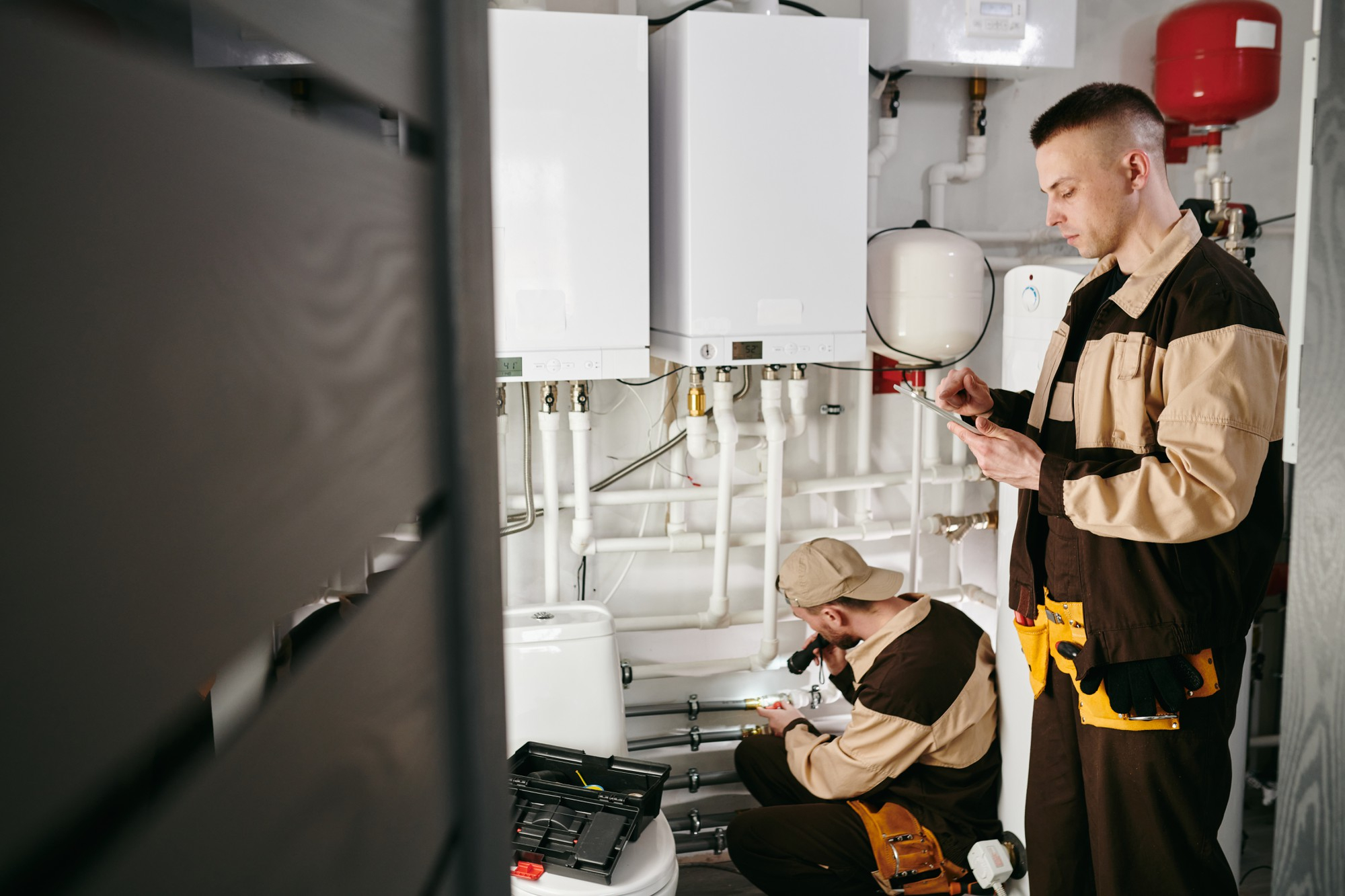 Two plumbers working on white boilers and pipes in a utility room, one using a flashlight and the other checking a tablet.