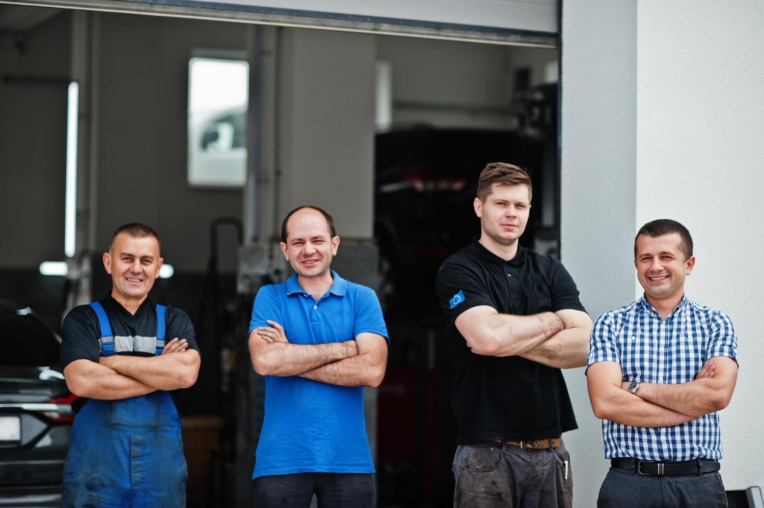 Four men standing with arms crossed in front of a garage entrance, two in work overalls and two in casual shirts.