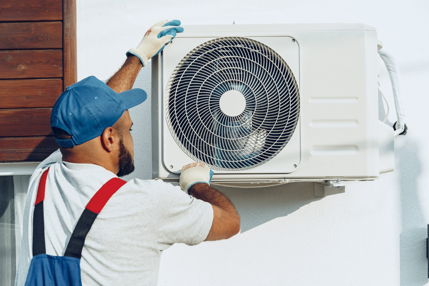 Technician wearing gloves and a blue cap installing or servicing an outdoor air conditioning unit on a white wall.