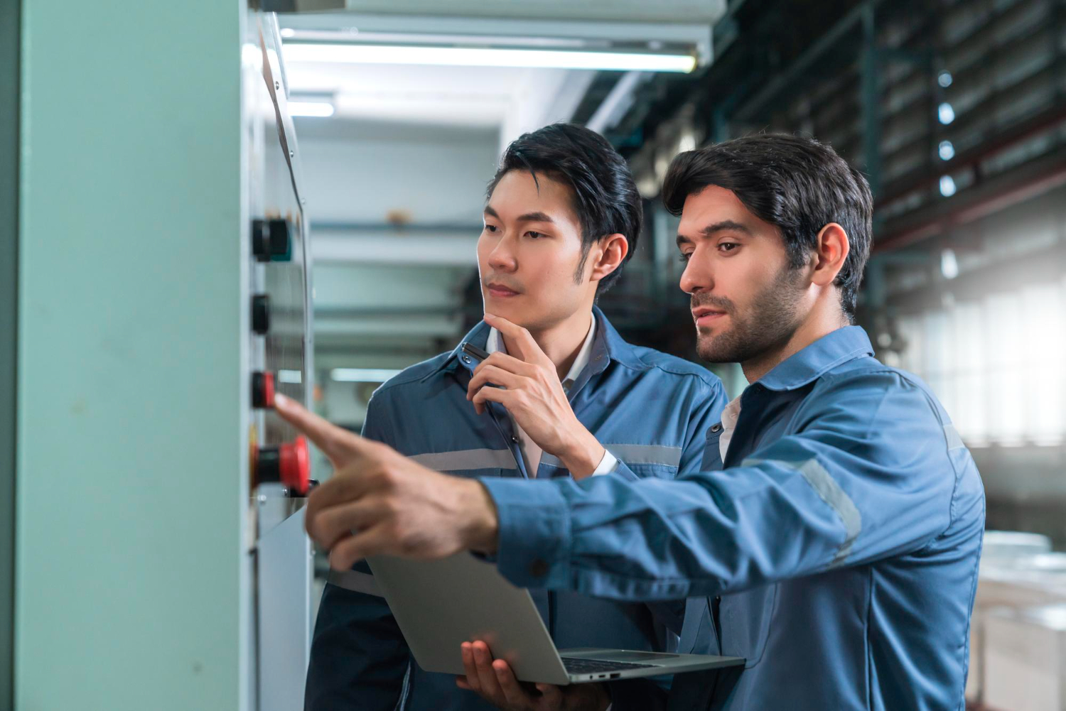 Two engineers in blue work uniforms operating control panel in an industrial setting, one holding a laptop.