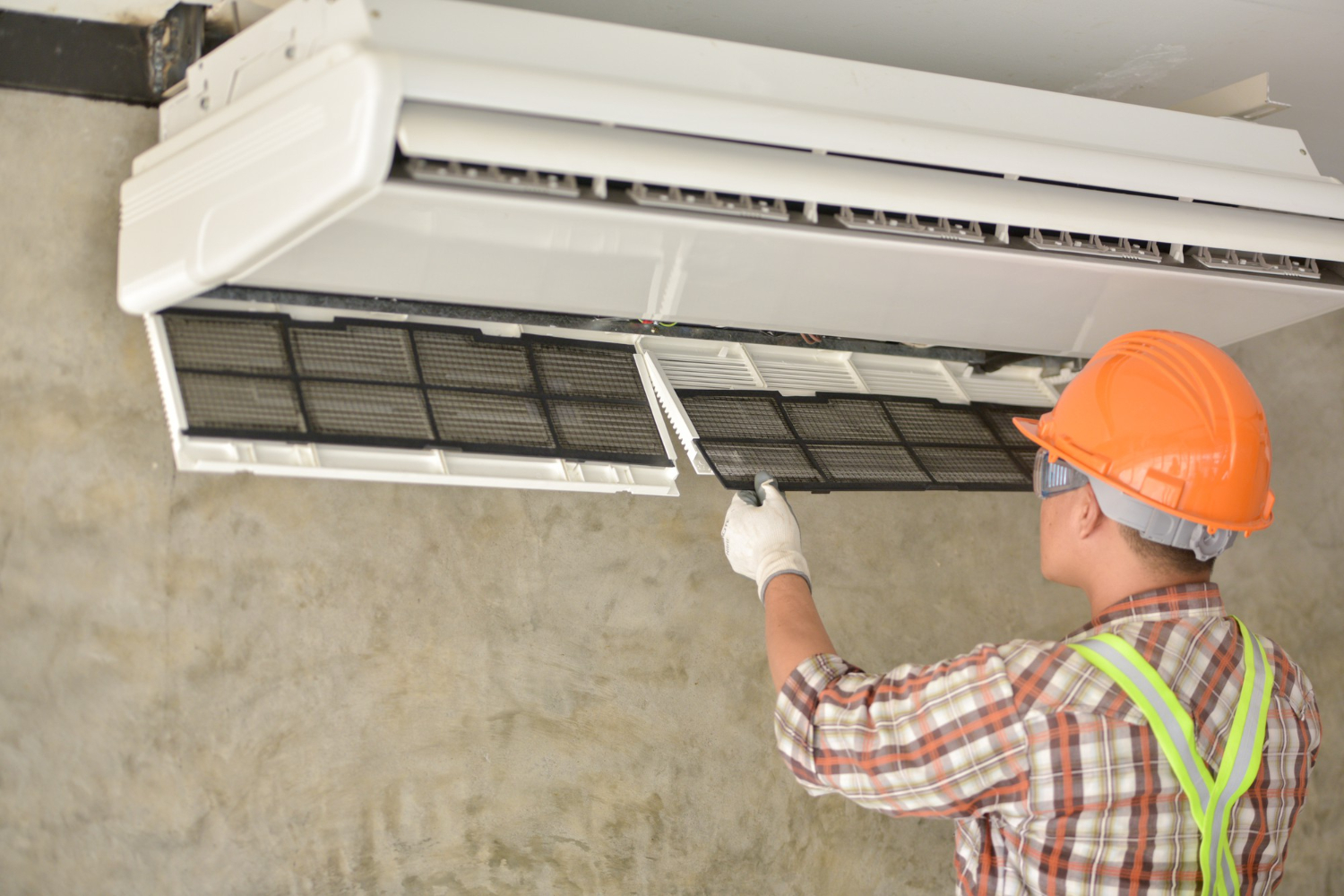 Technician wearing an orange hard hat and gloves removing a filter from a wall-mounted air conditioner.