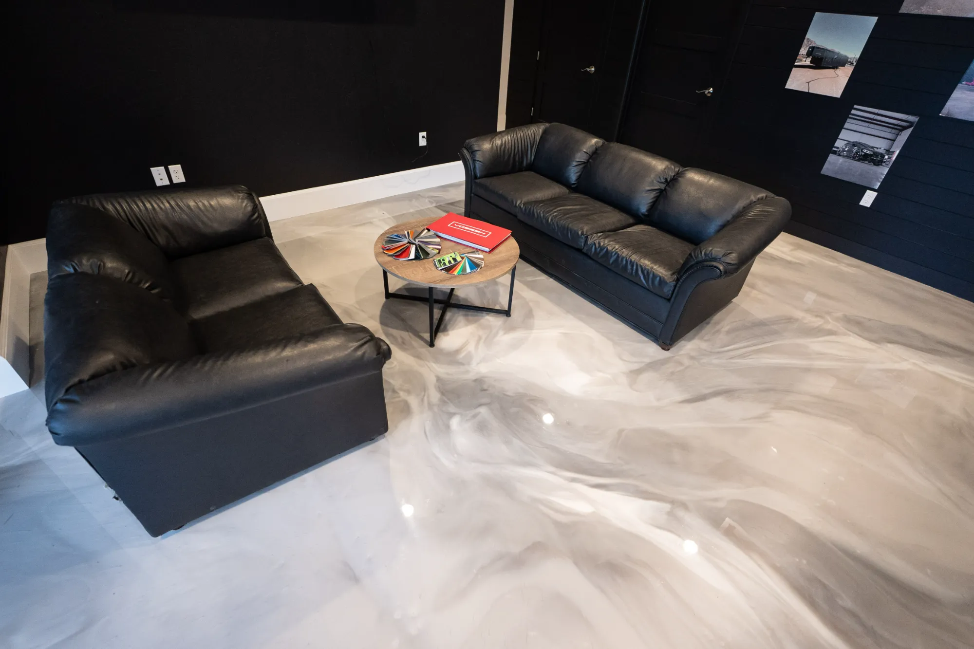 Modern room with two black leather sofas, a round wooden coffee table with color swatches and a red book, on a glossy marbled floor.