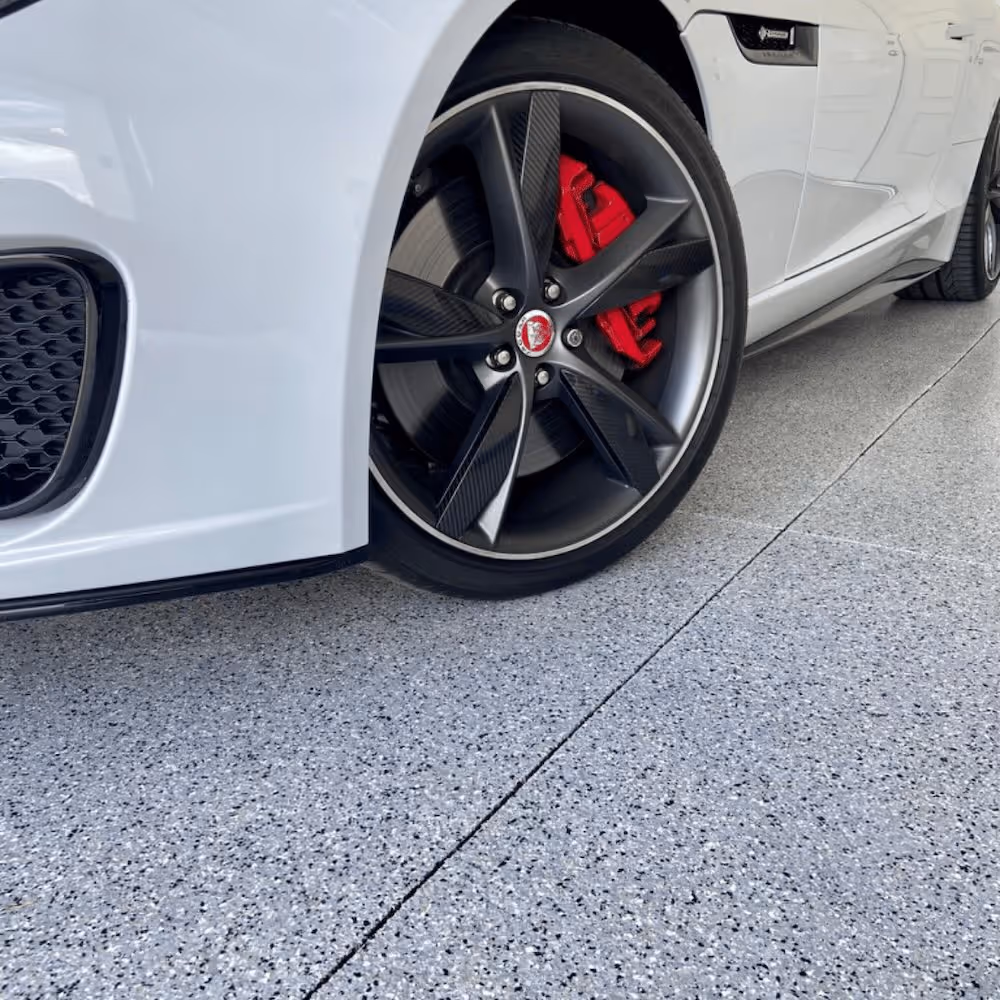 Close-up of a white car’s front wheel with black alloy rim and red brake caliper on speckled gray flooring.