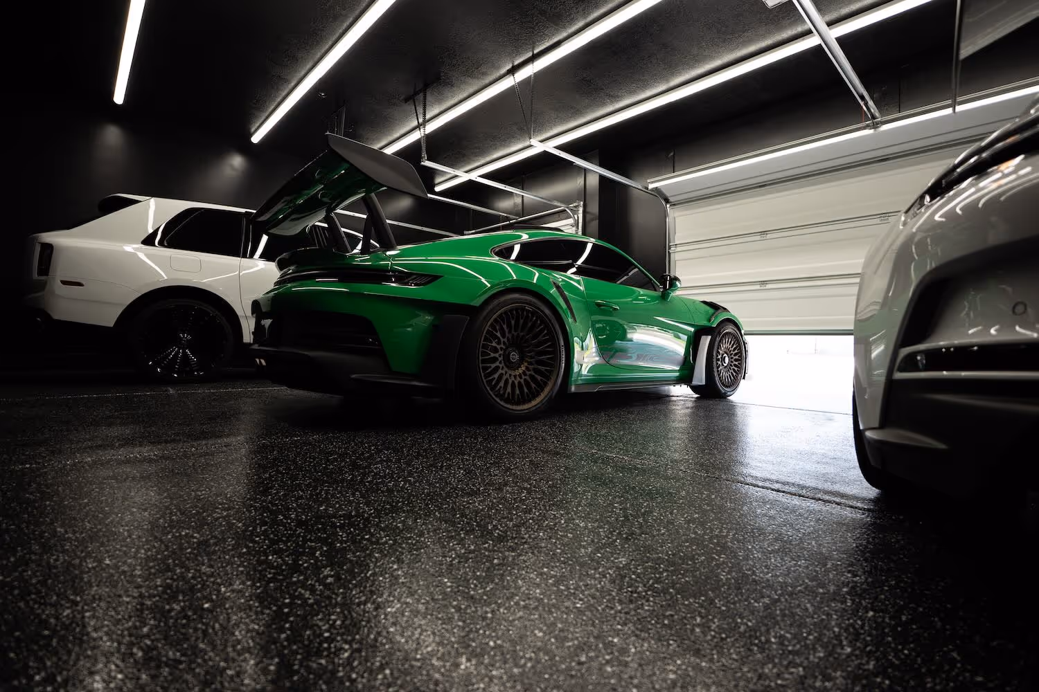 Green sports car with large rear wing parked in a dark garage next to a white SUV and another white vehicle.