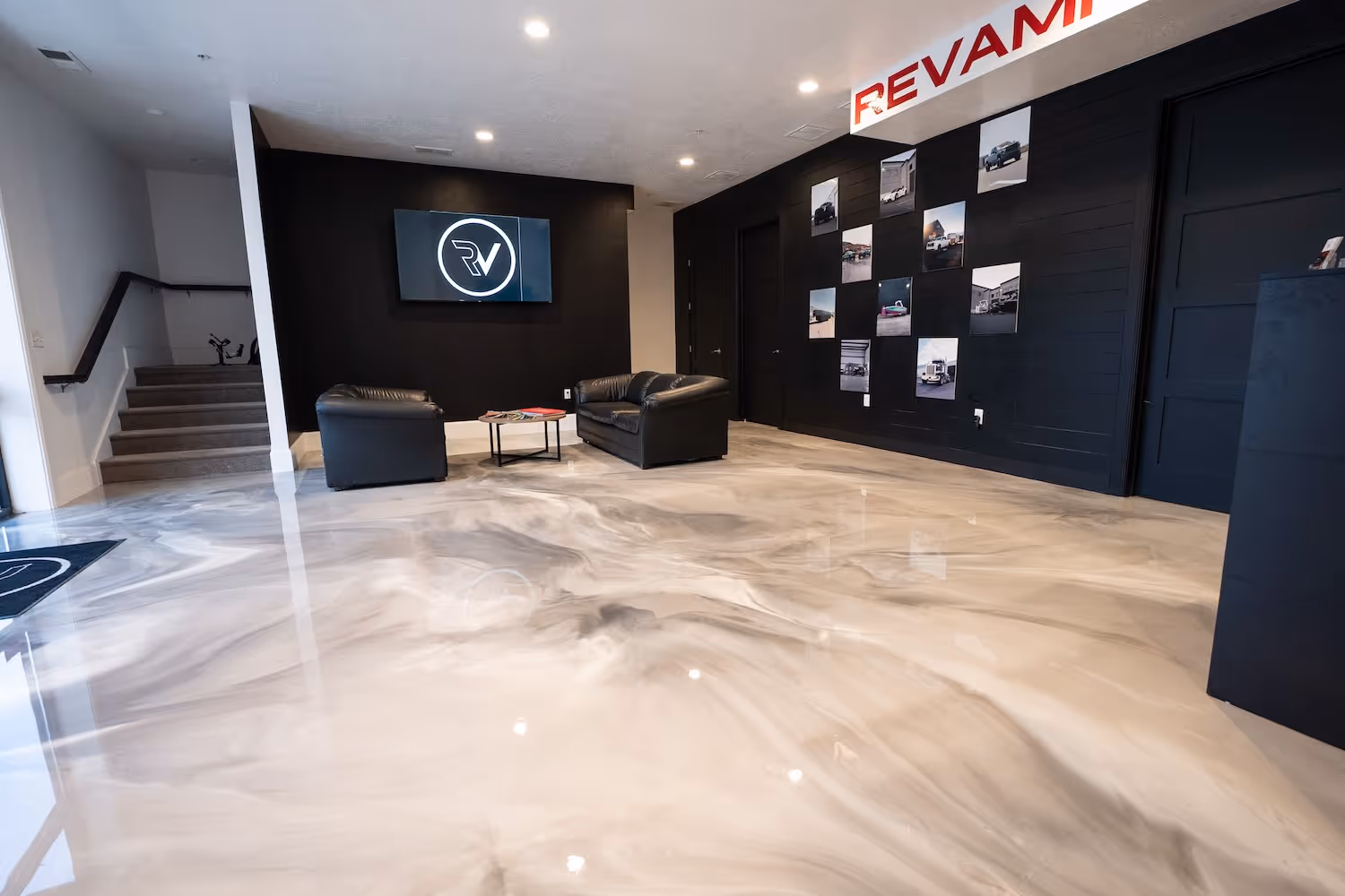 Modern office lobby with glossy marble floor, black leather sofas, a TV on a black wall, and photos displayed on a dark wall under a sign reading 'REVAMP'.