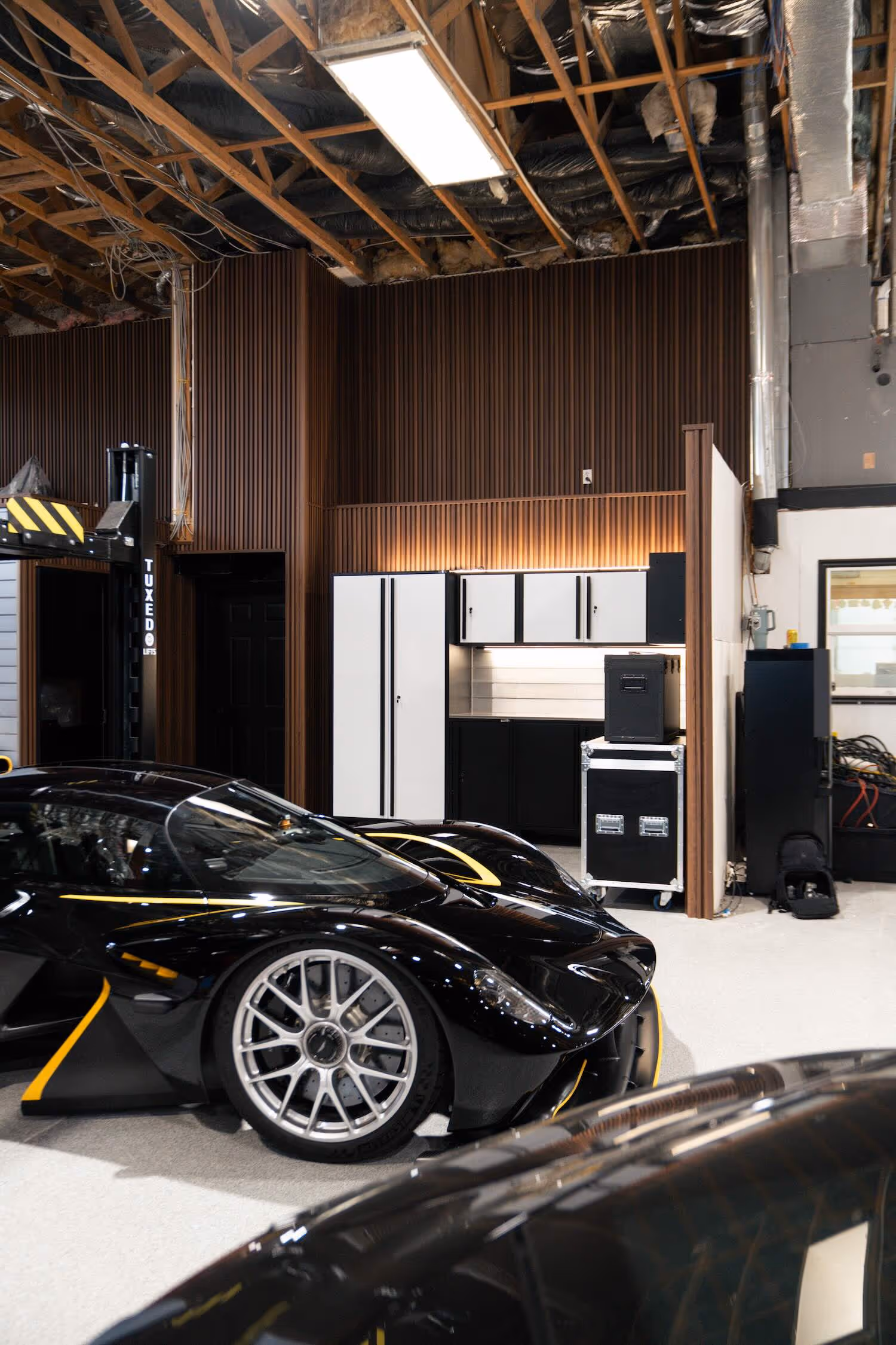 Black sports car with silver rims parked inside a garage with exposed ceiling and wooden panel walls.