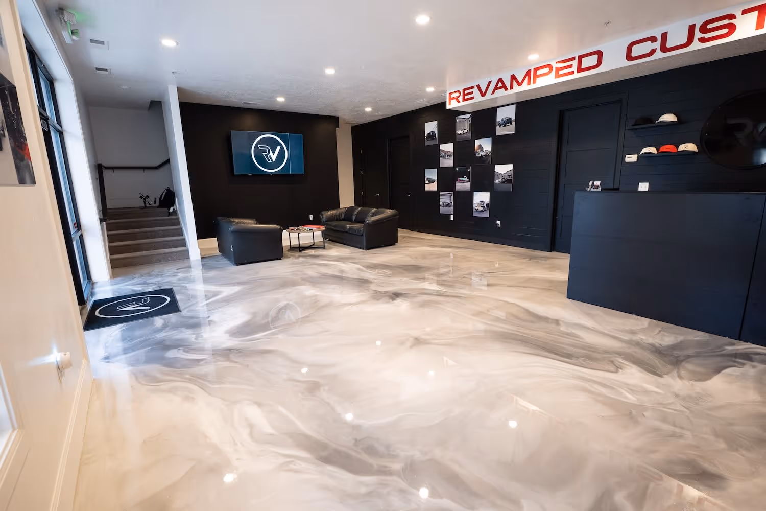 Modern office lobby with polished marble-look epoxy floor, black leather couches, TV with logo, and a photo wall display.