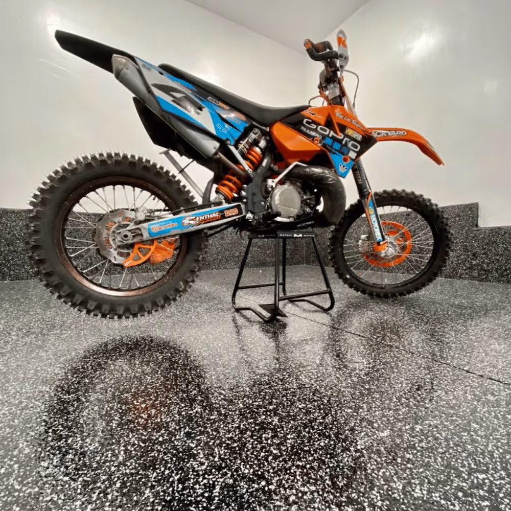 Orange and blue dirt bike with knobby tires and various stickers, displayed on a black stand indoors on a speckled floor.