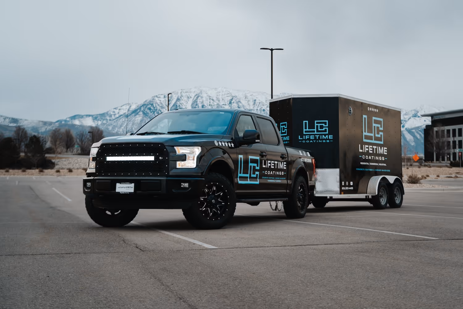 Black pickup truck with Lifetime Coatings branding parked with matching trailer in an empty lot with snowy mountains in the background.
