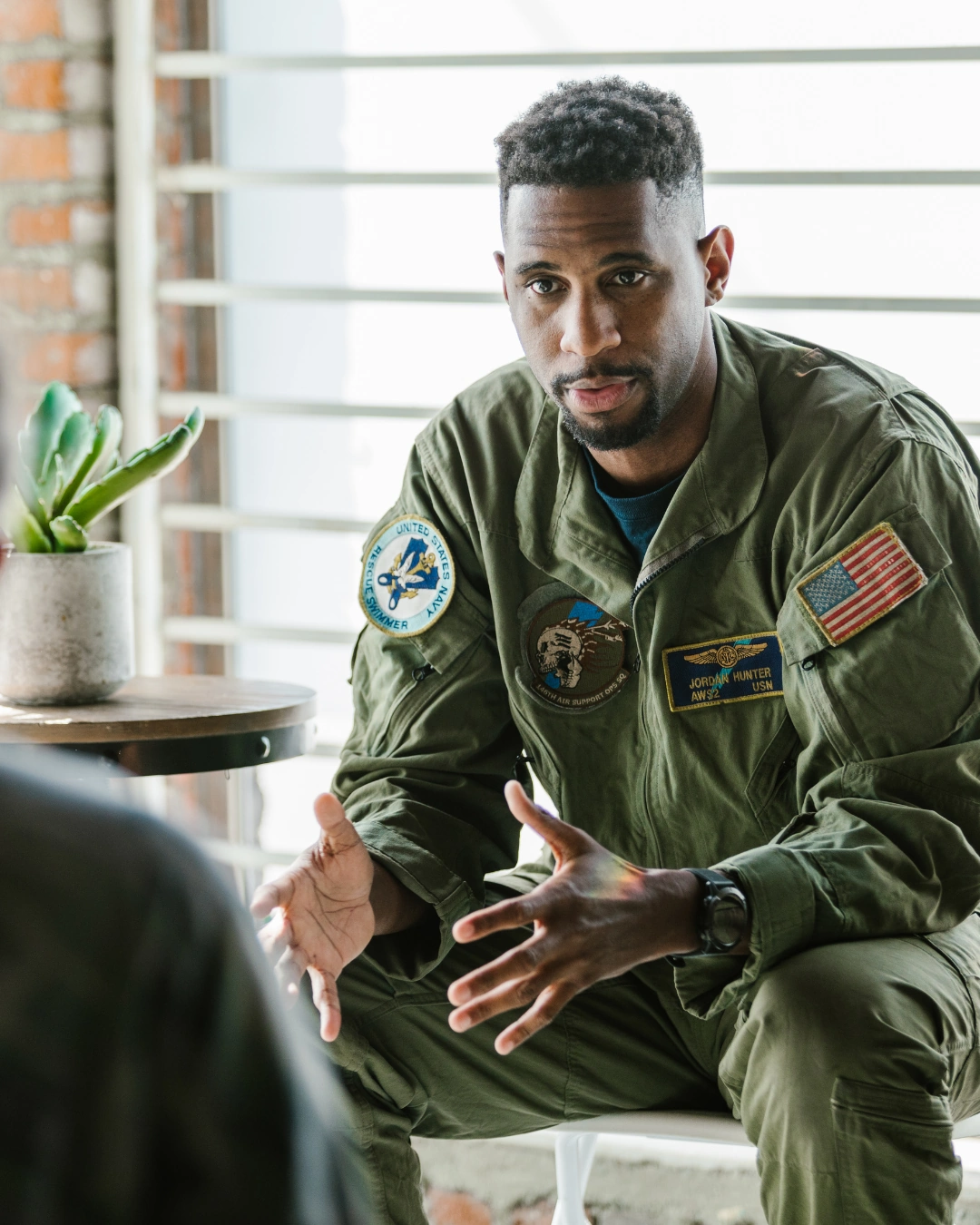 Man in U.S. Navy uniform with patches speaks, gesturing with hands indoors by a window and plant.