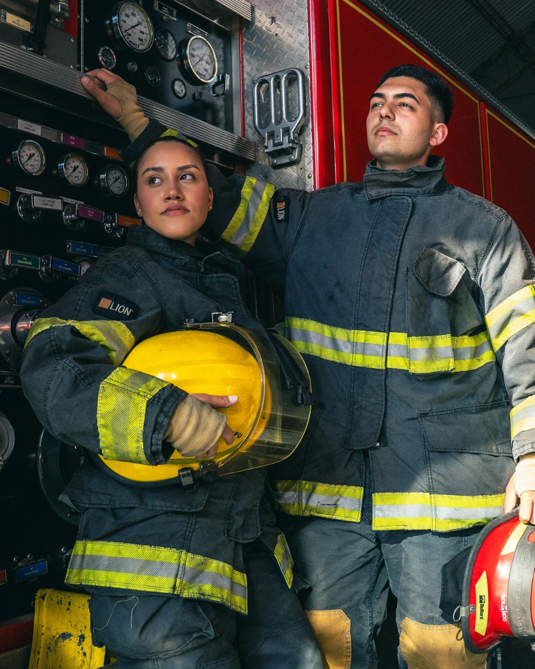 Two firefighters in gear, one holding a yellow helmet, standing by a fire truck panel with gauges.