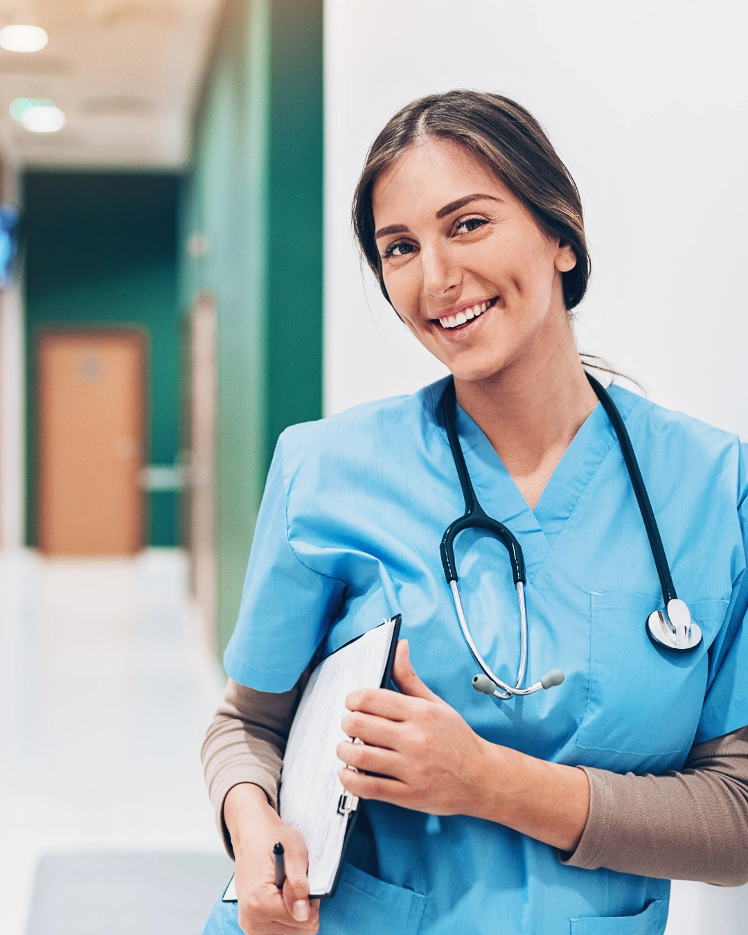 Smiling female nurse in blue scrubs holding clipboard with stethoscope around her neck.