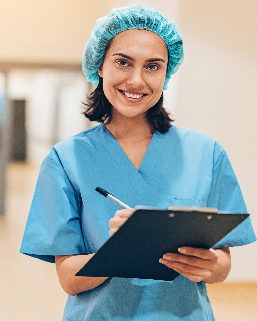 Smiling female healthcare worker in blue scrubs and hairnet holding a clipboard and pen.