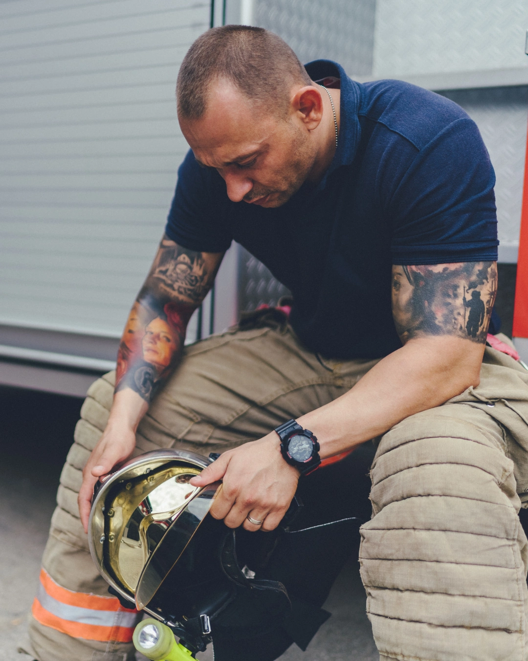 A firefighter with tattoos sits holding his helmet, looking down with a serious expression.