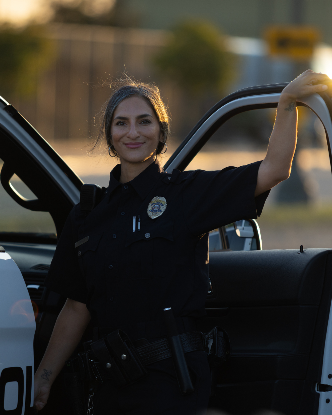 Smiling female police officer in uniform standing by an open police car door at sunset.