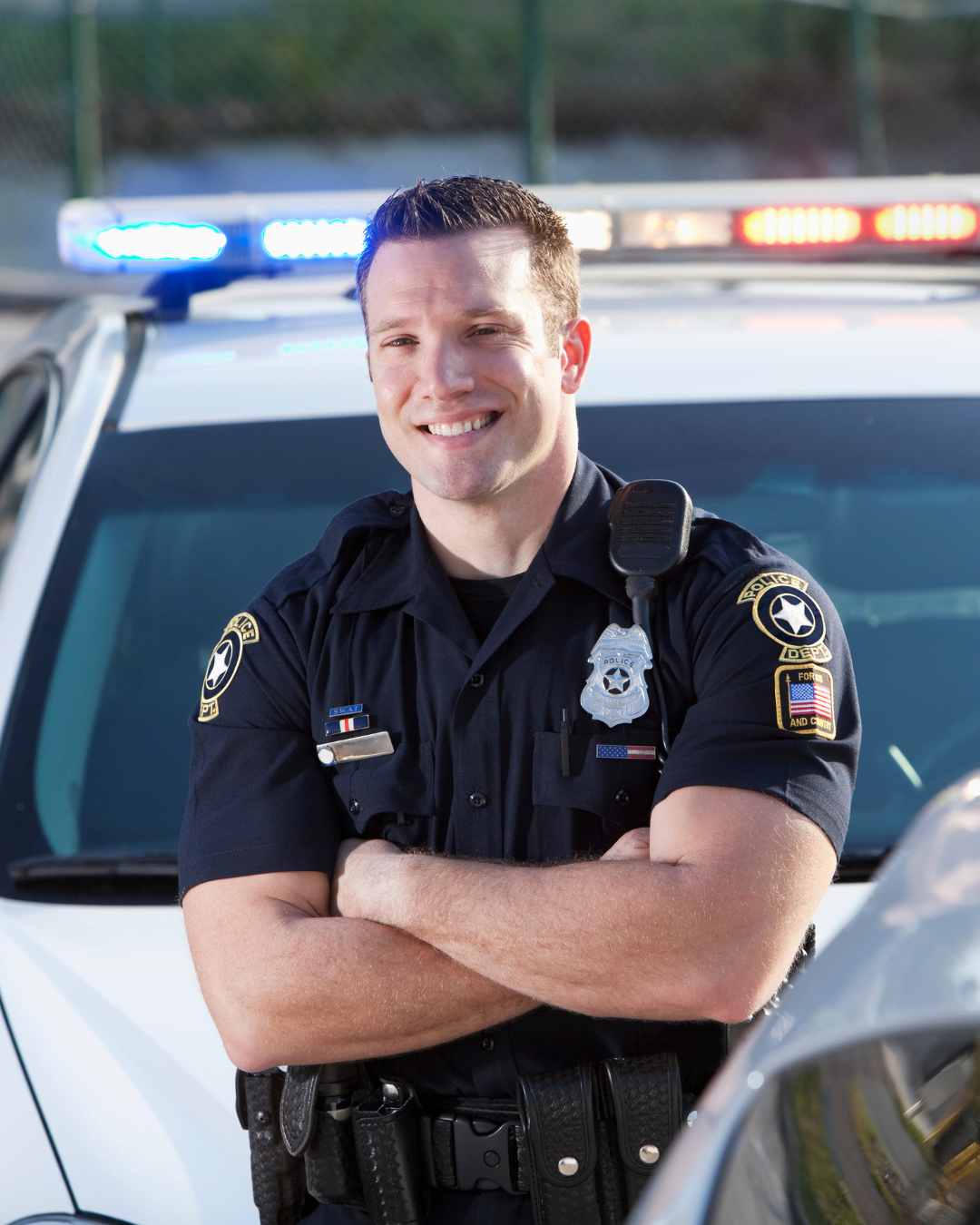 Smiling male police officer in uniform with arms crossed standing in front of police car with flashing lights.