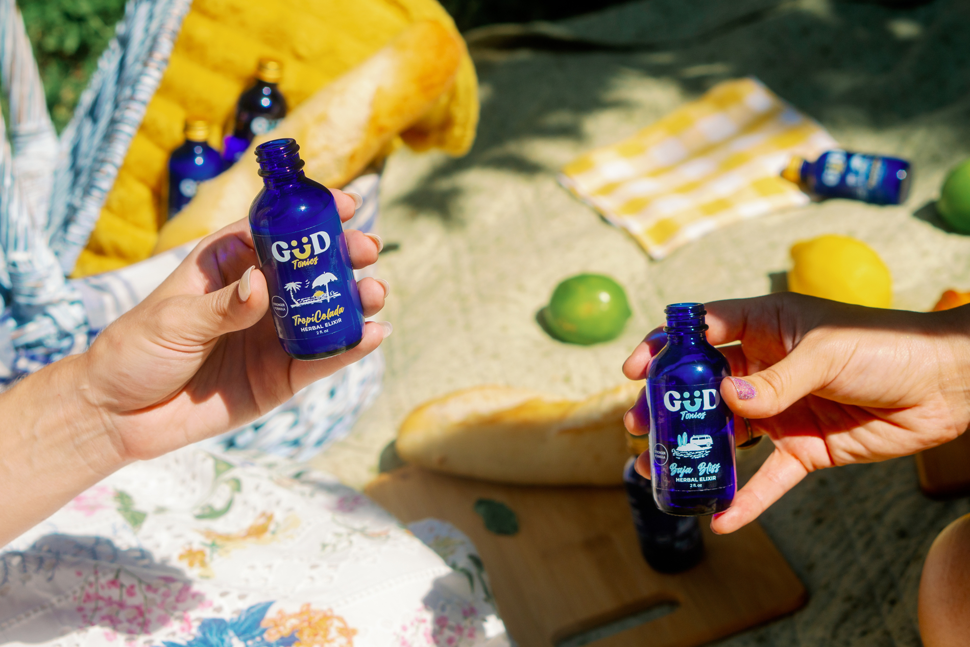 Two hands holding small blue bottles of GUD Tonics herbal elixirs on a sunny picnic blanket with bread, lemon, and lime in the background.