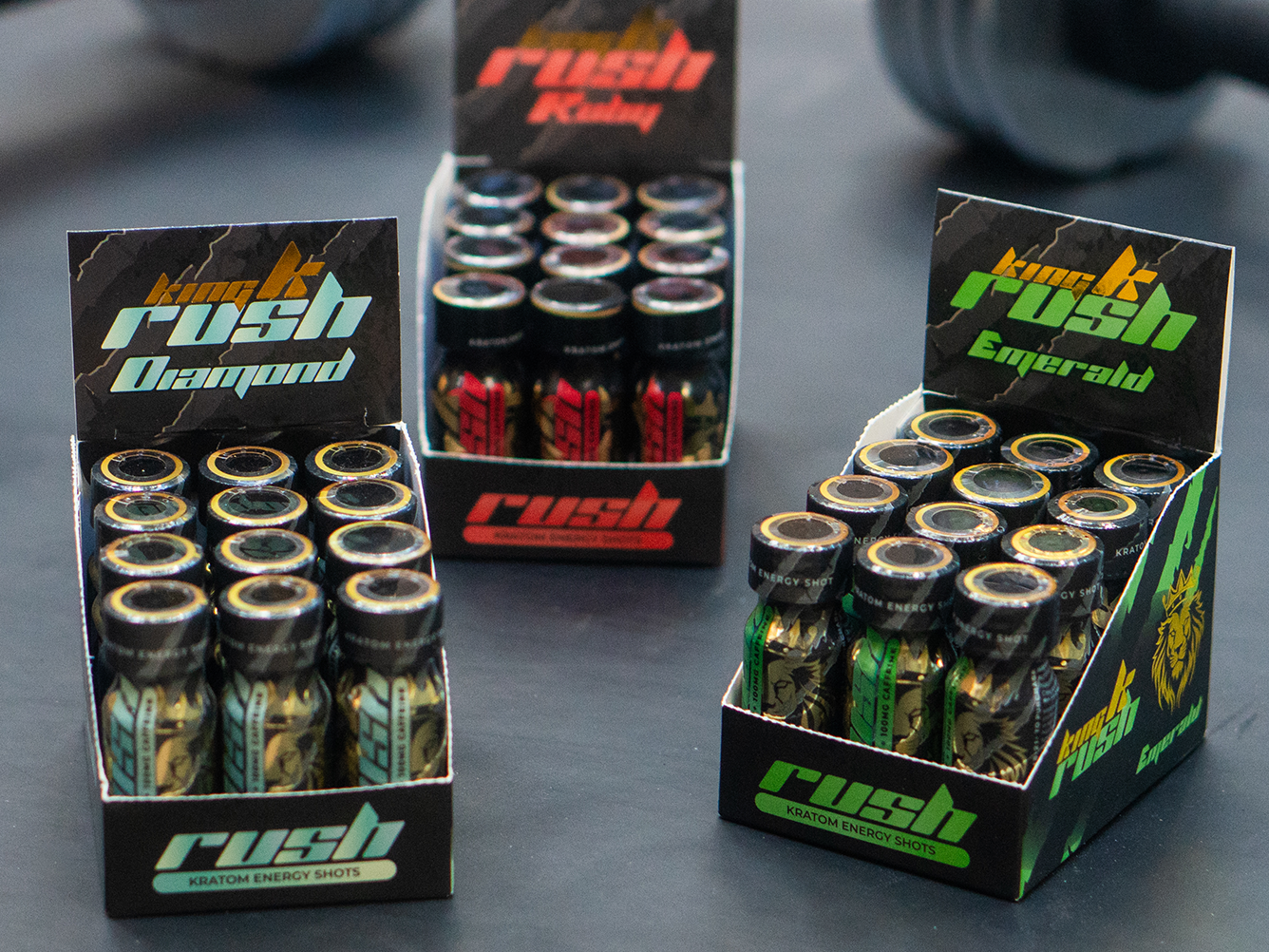 Three open boxes of King K Rush Kratom energy shots labeled Diamond, Ruby, and Emerald on a dark surface.