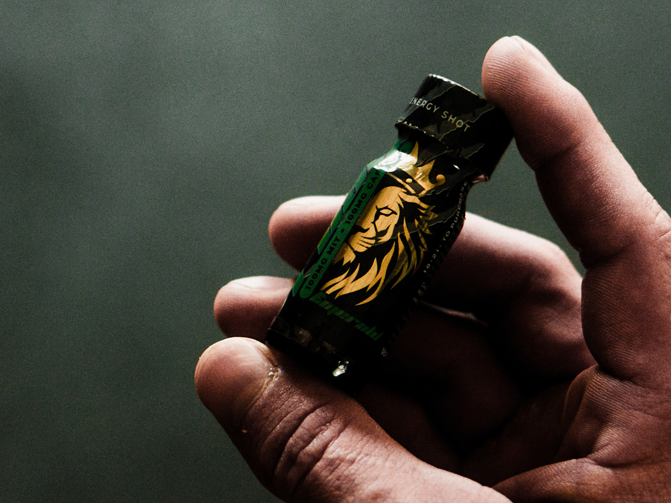 Hand holding a small dark green and black energy shot bottle with a gold lion logo.
