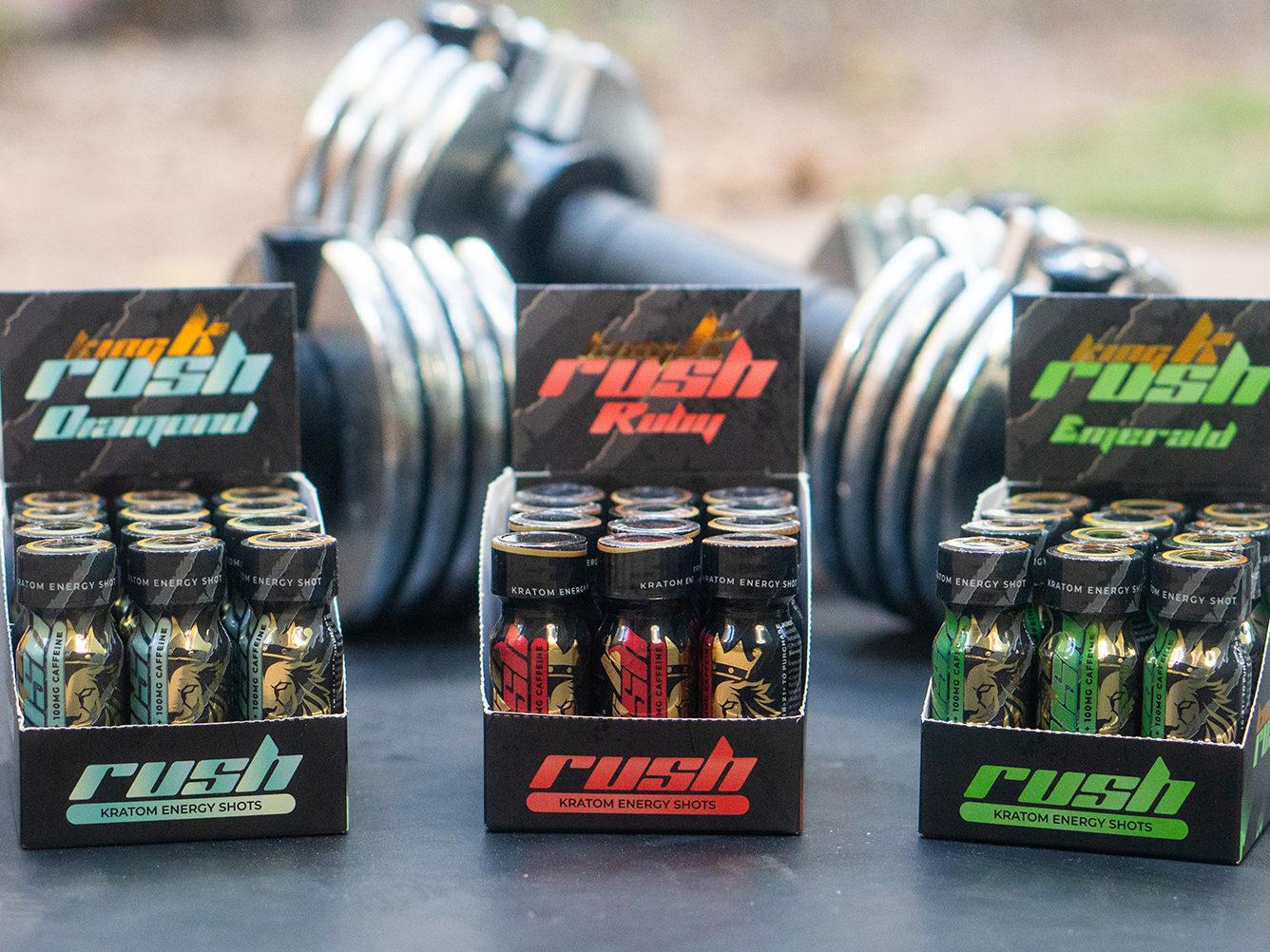 Three boxes of King K Rush Kratom Energy Shots in different flavors—Oceanaid, Ruby, and Emerald—with dumbbells blurred in the background.