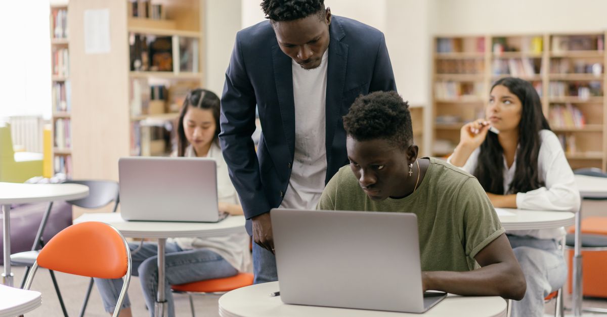 Studierende arbeiten mit Laptops in einer Bibliothek, eine Person berät sie
