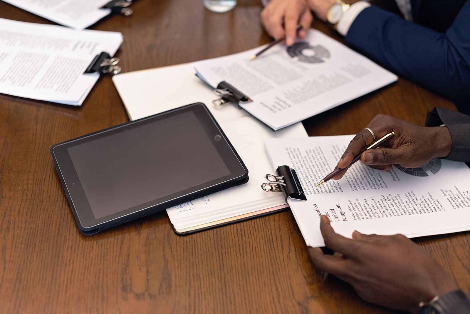 Hände mit Stift über Unterlagen und einem Tablet auf einem Besprechungstisch