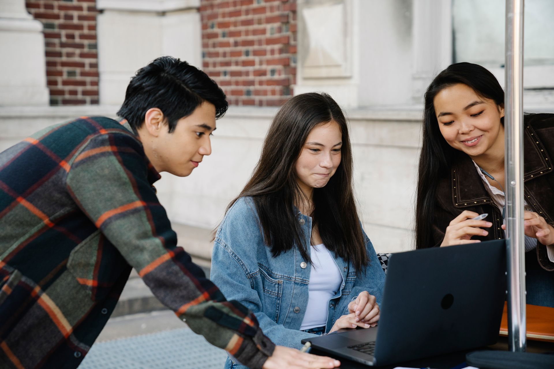 Drei Studierende schauen gemeinsam auf einen Laptop vor einem Hochschulgebäude